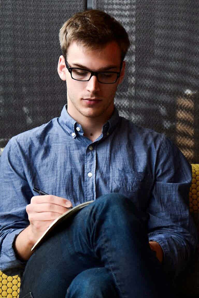 A young man in glasses writes in a notebook while sitting on a stylish couch indoors.