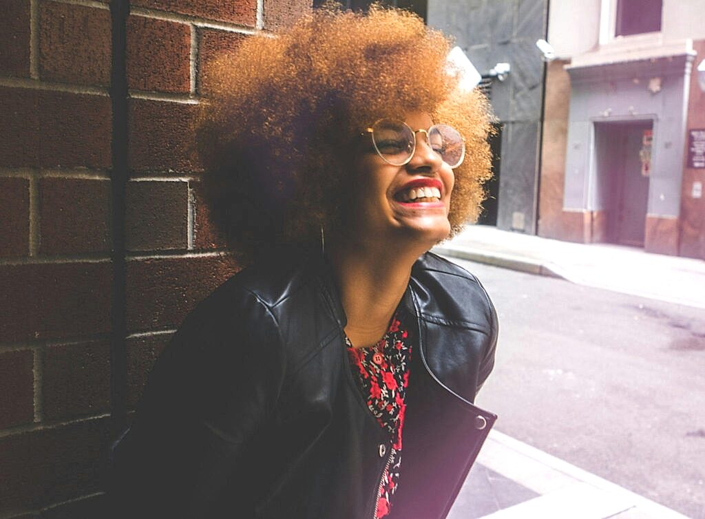 Joyful woman with afro hair and glasses smiles brightly outdoors in urban setting.